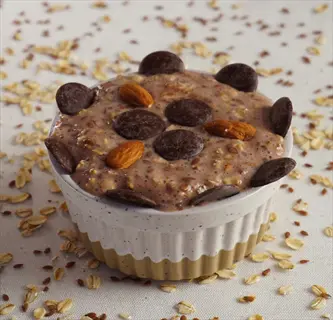 ramekin of oatmeal with chocolate pieces and almond toppings, surrounded by oats and flax seeds