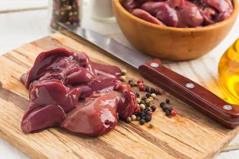 raw chicken liver pieces on a wooden cutting board with knife, peppercorns, and a bowl of additional liver in background