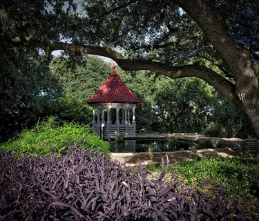 red-roofed gazebo in a botanical garden visit setting with a pond, lush greenery, and purple foliage under large tree branches