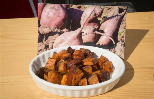 roasted sweet potatoes: golden-brown, seasoned cubes in white dish with farm-fresh raw sweet potato photo backdrop on wooden table