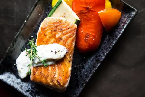 salmon vegetables plate: grilled salmon fillet with creamy dill sauce, served with roasted zucchini, yellow squash, and red bell peppers on a dark rectangular plate