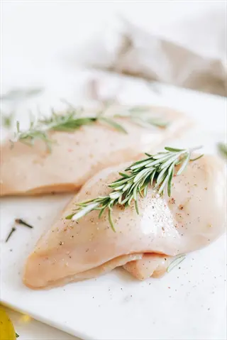 seasoned raw chicken breast pieces with rosemary sprigs and spices on a white cutting board, prepared for cooking