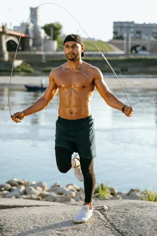 shirtless man jumping rope on a sunny day by a river with urban bridge and buildings in background