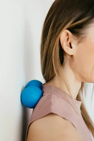 shoulder self massage: woman using blue massage balls against a wall for upper back and shoulder relief in a pink top