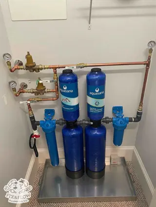 shower filter installation: aquasana whole-house water filtration system with blue canisters, copper pipes, pressure gauges, and valves on metal tray in utility closet