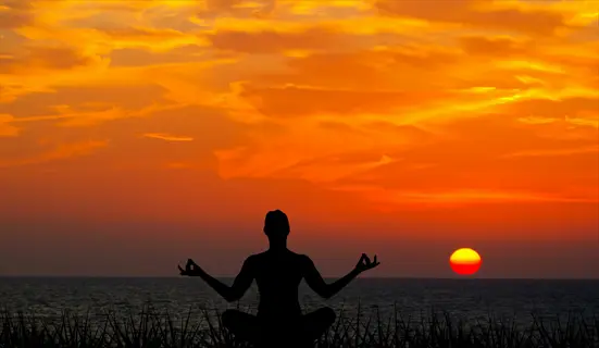 silhouette of a person meditating in a peaceful nature setting at ocean sunset - cross-legged pose with hands in mudra against vibrant orange sky