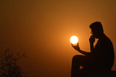 silhouette of a thoughtful person meditating at sunset, hand near face, sun positioned like a held orb in orange twilight sky with foliage