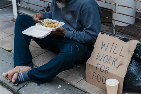 slow eating person sitting on urban sidewalk: barefoot individual in gray hoodie eats from takeout container beside 'will work for food' cardboard sign and paper cup