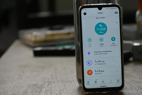 smartphone displaying fitbit fitness tracker app with daily stats (11,058 steps, 7.7 km) on a wooden table for progress tracking