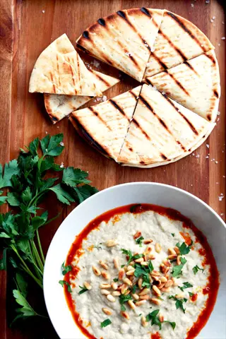 smoky eggplant dip (baba ganoush) in white bowl topped with pine nuts, parsley, and red pepper sauce, served with grilled flatbread on wooden board