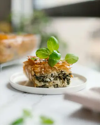 spanakopita feta pie slice with flaky filo layers, spinach and feta filling, garnished with fresh basil on white plate