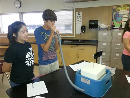 student demonstrating deep breathing into a scientific respirometer device during a lab class, with peers observing and lab materials visible