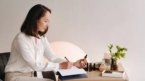 student practicing aromatherapy while studying: woman writes in notebook surrounded by essential oil bottles and diffuser in a serene, plant-filled room