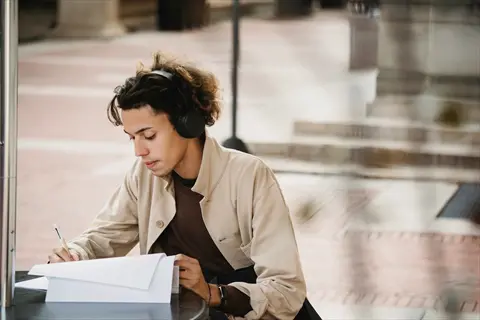 student wearing headphones studying outdoors while writing on papers at a table in an urban plaza setting