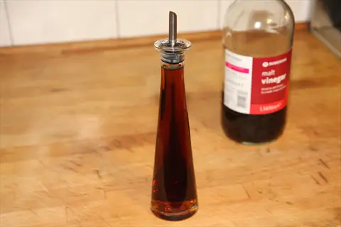 tall glass bottle vinegar with pour spout next to plastic malt vinegar bottle on wooden kitchen surface