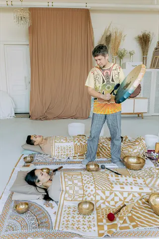 therapist conducting gong bath session with reclining participants under patterned blankets, surrounded by singing bowls in bright studio