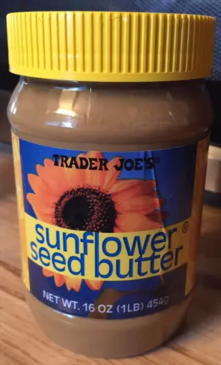 trader joe's sunflower seed spread jar with yellow lid, sunflower graphic, and visible text: 'trader joe's', 'sunflower seed butter', 'net wt. 16 oz (1lb 454g)' on wooden surface