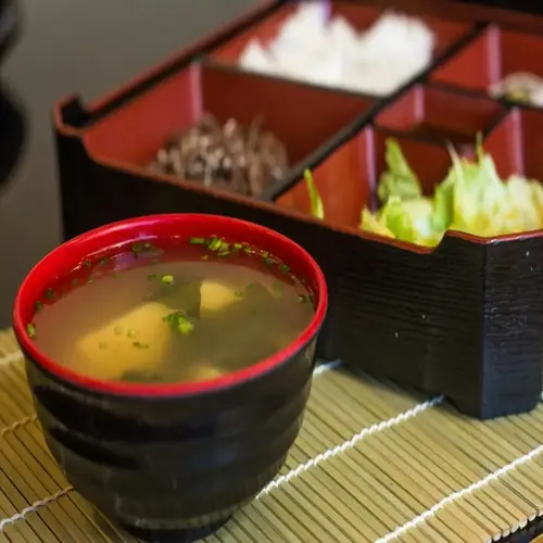 traditional miso soup bowl with tofu, seaweed and green onions beside japanese bento box with rice, meat, and vegetables on bamboo mat