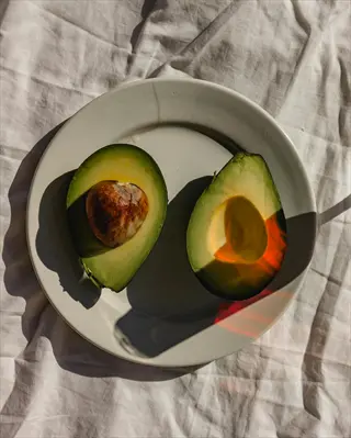 two avocado halves on white plate: one with pit, both showing creamy flesh. served as half portion under natural sunlight on wrinkled cloth