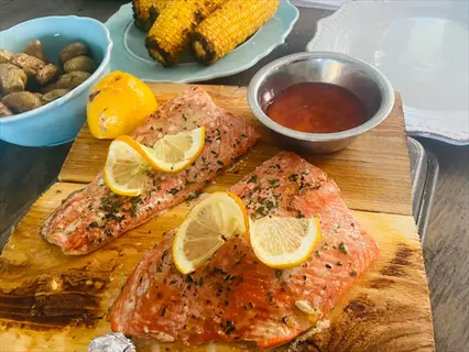 two cooked salmon portions on wooden board with lemon slices, herbs, and sides: roasted potatoes, corn, and dipping sauce. fatty fish fillets presented as a meal