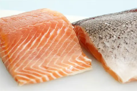 two fresh raw salmon fillets on a white surface - one skin-on with dark scales, one skin-off showing vibrant orange-pink flesh with white marbling