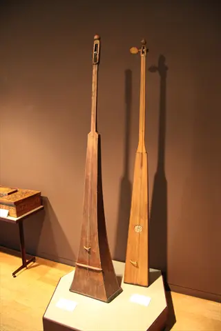 two large monochord instruments displayed on a pedestal in a museum gallery, with informational plaques and dramatic shadows against a taupe wall