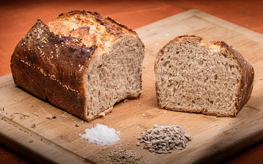 two loaves of whole grain bread (one sliced) on a wooden cutting board with sunflower seeds, salt crystals, and grains against an orange background