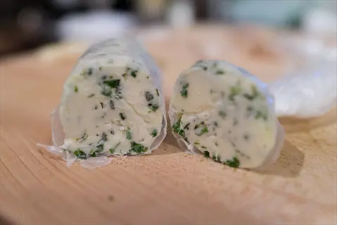 two logs of fresh herbal butter with visible herbs, wrapped in parchment on a wooden cutting board - herbal butters