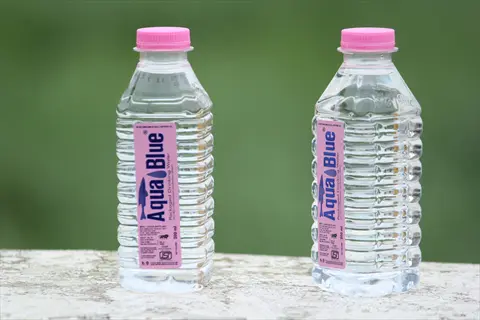 two mineral water bottles (aquablue brand) with pink caps on outdoor concrete surface against green foliage