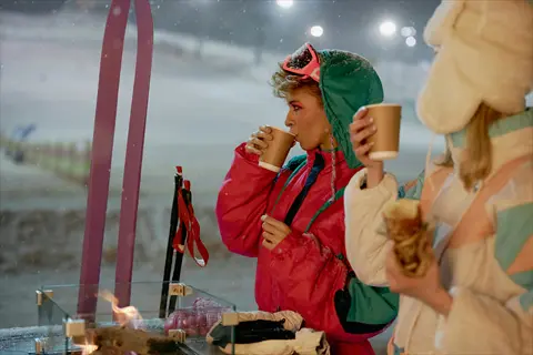 two people in winter gear drinking hot beverages before skiing at night with snowy slope in background