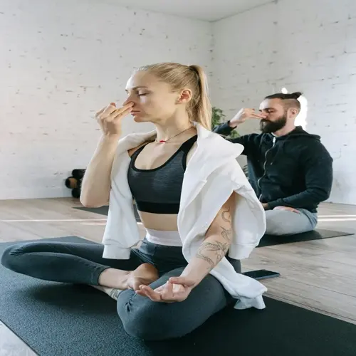 two people practicing breathing exercises in a yoga studio, woman in foreground and man in background seated on mats