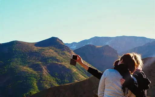 two people taking selfie with smartphone against mountainous hiking trail landscape under clear blue sky - travel adventure in nature