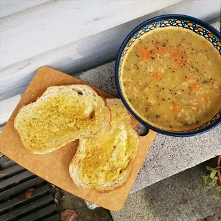 two slices of nutritional yeast bread on wooden cutting board and bowl of vegetable soup with carrots, outdoors on concrete slab near wooden structure