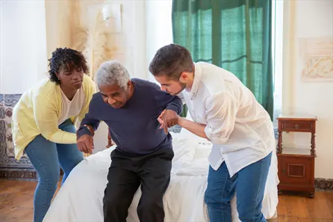 two volunteers helping elderly man rise from bed in sunlit room - embodying kind acts of service