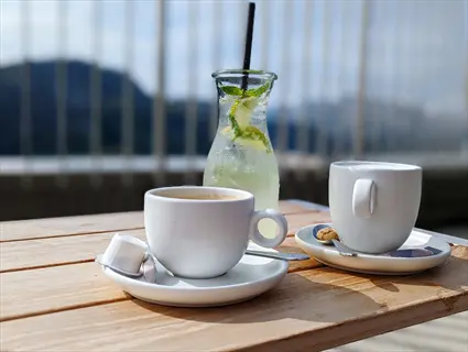 two white coffee cups, a glass of iced tea with lime and mint, and a water carafe on a wooden outdoor table with mountain scenery in background