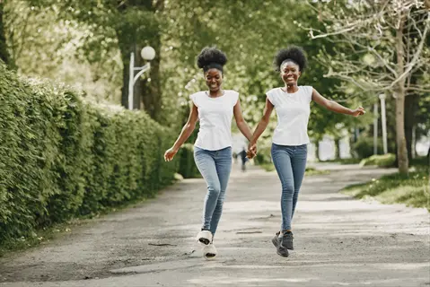 two women walking joyfully in a sunlit park, holding hands - embodying movement and wellness