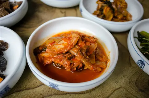 vibrant kimchi in white bowl with side dishes (banchan) on wooden table, showcasing fermented probiotic food