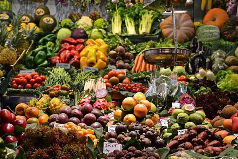 vibrant market stall overflowing with colorful fresh vegetables including bell peppers, carrots, pumpkins, and leafy greens, alongside labeled exotic fruits and produce