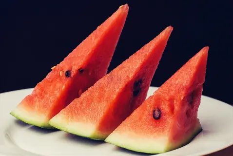 watermelon slices fresh: three triangular pieces with vibrant red flesh, black seeds, and green rind on white plate against dark background