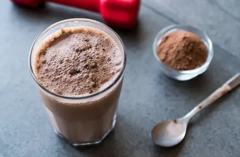 whey protein shake in a glass with a dumbbell, protein powder bowl, and spoon on a countertop. athlete nutrition for post-workout recovery