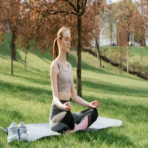 woman doing breathing exercise in a park: seated on yoga mat in lotus position with eyes closed, sneakers nearby on sunny grassy field with autumn trees