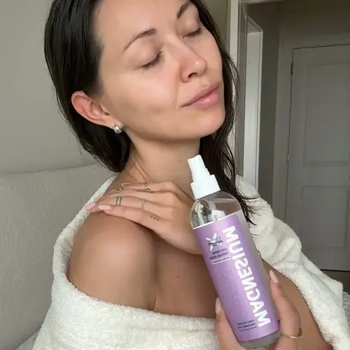 woman in white towel holding magnesium oil spray bottle with visible 'magnesium' text, demonstrating product use