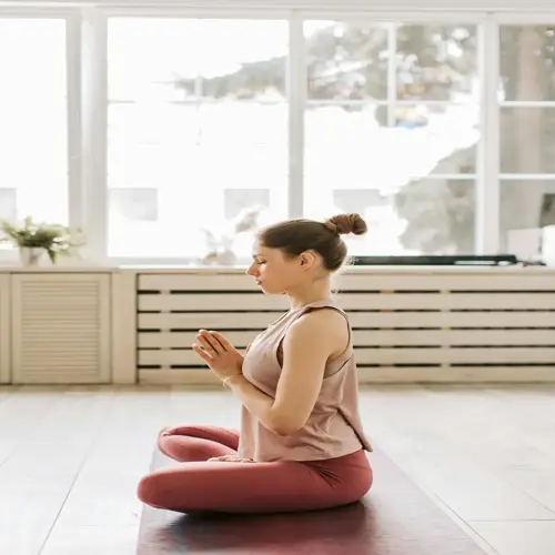 woman meditating at home in a yoga pose on a mat, hands in prayer position, bright room with large windows