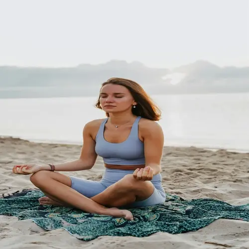 woman meditating in lotus position on a beach at sunrise, seated on a patterned cloth with ocean and morning sky