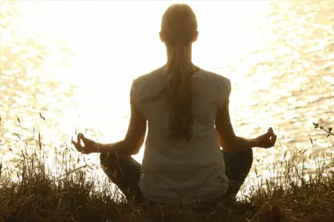 woman meditating in lotus position on grassy hill overlooking water at golden hour, embodying transcendental meditation practice