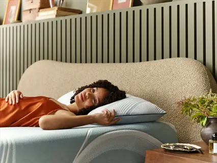 woman sleeping sideways on a bed in a modern bedroom, wearing an orange camisole and resting head on pillow against textured beige headboard with green vertical slat wall background