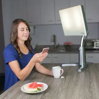 woman using carex day-light classic plus light therapy lamp at kitchen counter with phone, watermelon, and coffee mug (face visible, text present on lamp)