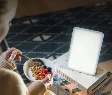 woman using lumie mini light therapy lamp during breakfast with cereal and fruit, next to a book titled 'breakfast'