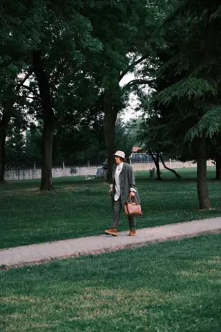 woman walking in a park along a paved path, wearing a hat and coat while carrying a brown bag, surrounded by tall trees and green grass